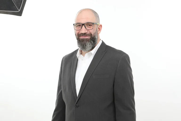 גבר מקצועי עם משקפיים וזקן בחליפה כהה וחולצה לבנה על רקע לבן